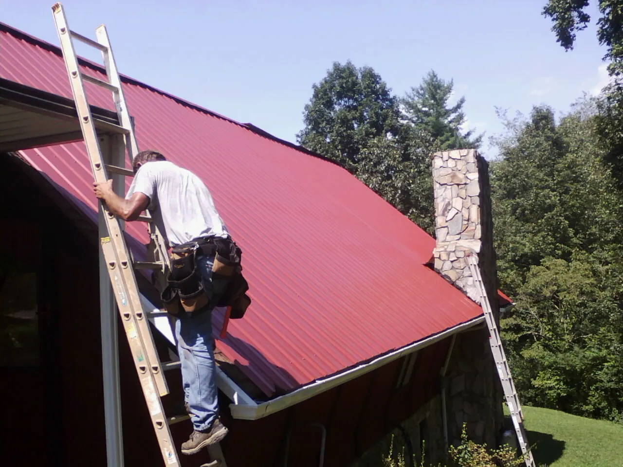 Roofer on a ladder beside a red metal roof and stone chimney — Soffit & Fascia Repair by Roofing Brothers in Garner